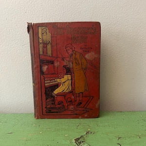 May include: Vintage book titled "The Organist's Baby" by Kathleen Knox. The cover is red with an illustration of a person and a child at an organ. The book is worn, with visible signs of age and use, and is set on a green surface.