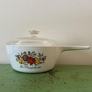 May include: White ceramic saucepan with a clear glass lid and a long handle. The saucepan features a colorful illustration of tomatoes, lemons, and other produce, with the text "La Marjolaine" below the illustration.