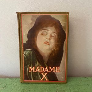 May include: Vintage book titled "MADAME X" featuring a portrait of a woman with dark hair and a somber expression. The book has a green cover with a red border and gold lettering. The book is resting on a green surface.