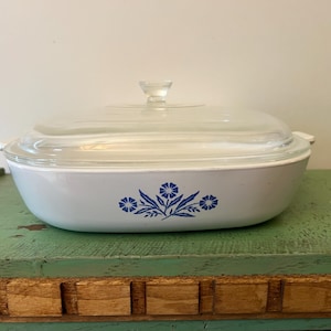 May include: A white casserole dish with a clear glass lid and a clear knob. The dish has a blue floral design on the front. The dish is sitting on a green surface.