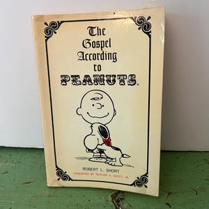 Peut inclure: Un livre vintage intitulé "The Gospel According to Peanuts". La couverture présente une illustration de dessin animé de Charlie Brown et Snoopy. Le livre a un fond crème avec du texte noir et des bordures décoratives. L'auteur est Robert L. Short.