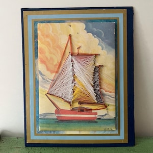 May include: A framed artwork featuring a sailboat crafted with thread. The boat is red and white, with white and yellow sails against a backdrop of a sunset sky. The artwork is framed in blue and gold.