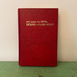 Pode incluir: Um livro vermelho com letras douradas que diz "WHY DOES THE DEVIL DESIRE TO DAMN YOU?". O livro está em pé sobre uma superfície verde. A capa do livro tem uma aparência texturizada.