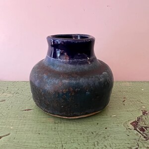 May include: A handcrafted ceramic vase with a rounded body and a dark blue glazed rim. The vase has a textured, mottled surface in shades of blue and green. The vase sits on a light-colored base.
