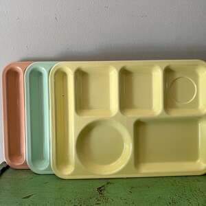 May include: Three vintage cafeteria trays in pastel colours. The trays are rectangular with multiple compartments for food. One is peach, one is mint green, and the third is yellow.