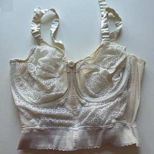 May include: Ivory-colored vintage bra with lace detailing on the cups and straps. The bra has a structured design with a front closure and adjustable straps. The fabric appears to be a combination of satin and lace.