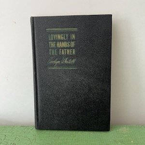May include: A black hardcover book titled "LOVINGLY IN THE HANDS OF THE FATHER" by Evelyn Whitell. The book has a textured cover and the title is in green and white lettering. The book is standing upright on a green surface.