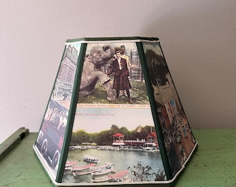 May include: A vintage lampshade with a green trim and white edging. The shade features several postcard images, including a baby elephant with a keeper, a lake with boats, and a city scene. The text on the shade reads "Baby Elephant; showing keeper and his daughter; Central Park, New York."
