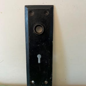 Puede incluir: Placa de puerta rectangular negra con cerradura y abertura circular para el pomo. La placa tiene cuatro orificios para tornillos y un acabado ligeramente desgastado, lo que sugiere una estética antigua o vintage.