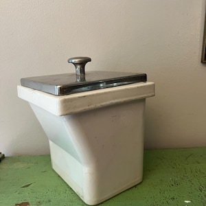 May include: A white, angled container with a stainless steel lid and a rounded handle. The container sits on a distressed green surface, suggesting a vintage or antique aesthetic. The lid is rectangular and the handle is centered.