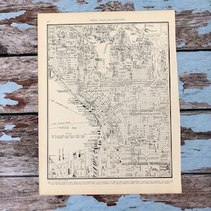 May include: A vintage map of Seattle, Washington, showing the city's streets, waterways, and surrounding areas. The map is printed in black and white on cream-coloured paper. The text "WORLD ATLAS AND GAZETTEER" is printed at the top of the map.