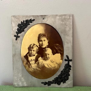Puede incluir: Retrato familiar antiguo en tonos sepia en un marco ovalado. El marco es rectangular con un acabado gris claro desgastado y detalles florales negros. El retrato muestra a una mujer y dos niños.
