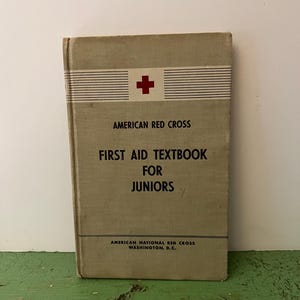 May include: A vintage light gray hardcover book titled American Red Cross First Aid Textbook for Juniors. The cover features a red cross symbol, horizontal black lines, and the text American National Red Cross Washington, D.C. at the bottom.