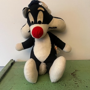 Può includere: Un peluche vintage di Silvestro il Gatto, con un corpo nero e bianco, un naso rosso e grandi occhi espressivi. Il giocattolo è seduto, con le braccia e le gambe distese. C'è un'etichetta attaccata.