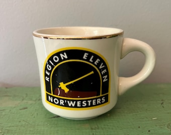 Puede incluir: Una pequeña taza de cerámica blanquecina con borde y asa dorados. La taza presenta un emblema circular con el texto "REGION ELEVEN NOR'WESTERS" en negro y amarillo. El emblema incluye un gráfico de una herramienta amarilla.