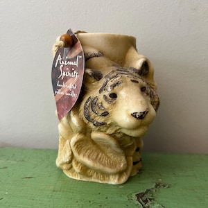 May include: A beige pillar candle shaped like a tiger, with black stripes and facial details. The candle has a tag that reads "Animal Spirits, handcrafted pillar candles."