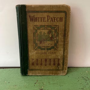 May include: Vintage hardcover book titled "White Patch" by Angelo Patri. The cover is tan with green and brown accents, featuring an illustration and text. The spine is dark green. The book is standing on a green surface.