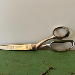 Puede incluir: Tijeras Wiss Professional Model AA plateadas. Las tijeras tienen grandes orificios para los dedos y una hoja curva. El texto "Wiss Professional Model AA" está impreso en la hoja. Las tijeras están sobre una superficie verde.