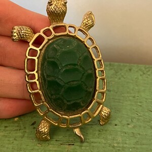 May include: Vintage gold-tone turtle pendant with a textured green center. The turtle's shell is an oval shape, and the pendant has a loop at the top for hanging. The turtle's head, feet, and tail are detailed.