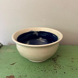 May include: A ceramic bowl with a cream-colored exterior and a deep blue interior. The bowl has a rounded shape with a slightly flared rim. The exterior has a crackled texture, and the bowl sits on a green surface.