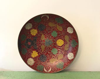 May include: A circular decorative bowl featuring a deep red base and detailed floral patterns in multiple colours. The design incorporates yellow, green, blue, and white flowers, alongside paisley and crescent moon motifs. The bowl has a dark rim.
