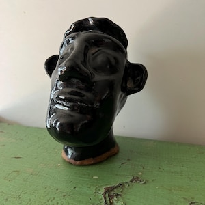 May include: A black ceramic head sculpture with a glossy finish. The sculpture depicts a human face with a beret. The base has a brown rim. The sculpture is on a green surface.