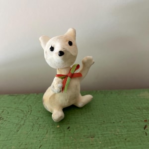 May include: A vintage white dog figurine, seated with black eyes and nose. It has a red and green ribbon around its neck. The figurine is on a green surface.