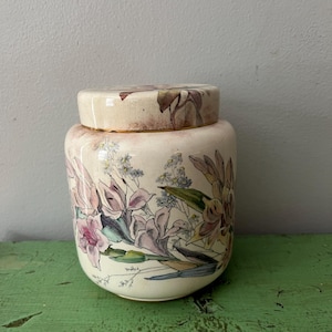 Puede incluir: Un tarro de cerámica color crema con tapa, adornado con un diseño floral en tonos rosa, morado y verde. El tarro tiene forma redondeada y está sobre una superficie verde. La tapa presenta un patrón floral a juego.