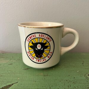 Peut inclure: Mug en céramique crème avec un bord doré et une anse incurvée. Le mug présente un emblème circulaire avec une tête de taureau noire, le chiffre 12 et les mots "SIEMPRE ADELANTE" et "REGION TWELVE B.S.A." en rouge et noir.
