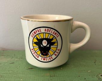 Puede incluir: Taza de cerámica color crema con borde dorado y asa curvada. La taza presenta un emblema circular con una cabeza de toro negra, el número 12 y las palabras "SIEMPRE ADELANTE" y "REGION TWELVE B.S.A." en rojo y negro.