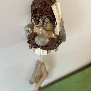 Könnte beinhalten: Eine skurrile Keramikskulptur eines Mannes mit braunem Haar, Brille und Schnurrbart, der ein Buch hält. Die Skulptur hat ein einzigartiges, handgefertigtes Aussehen mit hängenden Beinen. Die Farbpalette umfasst Brauntöne, Beige und Grau.