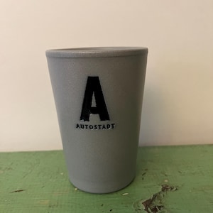 May include: A gray tumbler with a black letter "A" and the word "AUTOSTADT" printed below it. The tumbler has a slightly tapered shape and a matte finish. The background is a light green surface.
