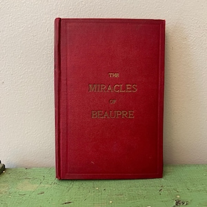 Pode incluir: Um livro de capa dura vermelho com o título "THE MIRACLES OF BEAUPRE" em letras douradas. O livro tem uma capa texturizada e está em pé. O fundo é uma parede de cor clara e uma superfície verde.