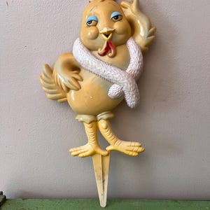 May include: A whimsical, yellow chicken figurine with a white scarf and a red comb. The chicken has blue eye shadow and a red wattle. It is mounted on a stake, likely for garden or yard decor.