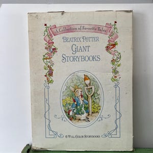 Puede incluir: Un juego de cajas vintage de Beatrix Potter Giant Storybooks. La caja presenta el título "A Collection of Favorite Tales" en escritura rosa, con ilustraciones de Peter Rabbit y otros personajes. La caja es beige con una base verde.