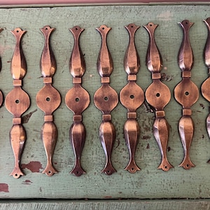 Könnte beinhalten: Eine Sammlung von zehn antiken Schrankgriffen in Bronzetönen. Jeder Griff hat ein dekoratives Design mit einer abgerundeten Mitte, einem Loch und einem stilisierten oberen und unteren Teil. Die Griffe sind auf einer verwitterten grünen Oberfläche angeordnet.