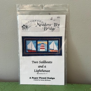Op de afbeelding: Een papieren patchwork ontwerp getiteld "Two Sailboats and a Lighthouse" van Meadow Lily Bridge. Het ontwerp toont twee zeilboten en een vuurtoren. De geschatte afmeting is 56 cm x 28 cm. Het ontwerp zit in een doorzichtige plastic hoes.