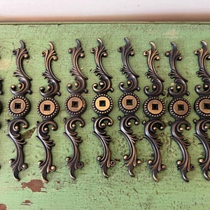 May include: A collection of ornate, bronze-toned drawer pulls. Each pull features a scrolling design with a central circular element containing a square inset. The pulls are arranged in rows, showcasing their decorative details and vintage aesthetic.