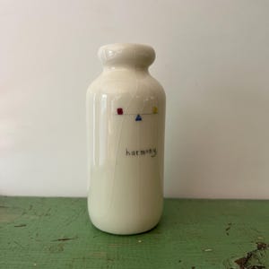 Puede incluir: Una pequeña botella de cerámica color blanco hueso con un acabado liso y brillante. El frente presenta un delicado dibujo lineal de una balanza con un cuadrado rojo, un triángulo azul y un cuadrado amarillo. La palabra harmony está impresa en negro.