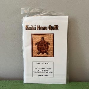 Pode incluir: Um kit de colcha Keiki Honu embalado. O design da colcha apresenta uma tartaruga em uma moldura marrom. A embalagem é selada em plástico transparente. O tamanho é de 71 cm x 76 cm.