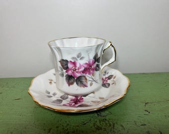 May include: A white teacup and saucer set with a floral design. The cup and saucer are decorated with pink flowers, gray leaves, and gold trim. The teacup has a curved handle.