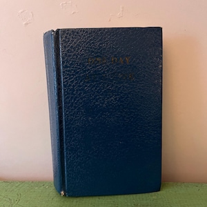 Puede incluir: Un libro pequeño, azul oscuro con letras doradas en la portada. El libro tiene una cubierta texturizada y un lomo. El libro está de pie sobre una superficie verde.