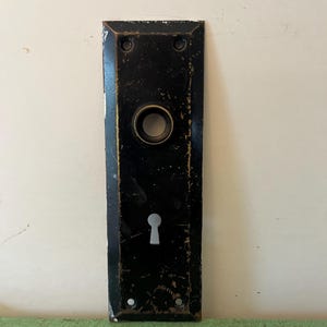 Puede incluir: Placa de puerta rectangular negra antigua con cerradura y abertura circular para el pomo. La placa tiene un acabado envejecido con detalles dorados en los bordes y alrededor de la abertura circular.