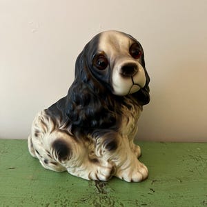 May include: A ceramic figurine of a Cocker Spaniel dog, featuring a black and white coat with reddish-brown eyes. The dog is in a seated position, with its head tilted slightly. The figurine is on a green surface.