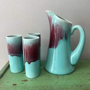 May include: A turquoise ceramic pitcher and three matching tumblers. The pitcher has a large handle and a maroon glaze that drips down the turquoise body. The tumblers have a similar design, with maroon glaze at the top and turquoise at the bottom. The set is likely vintage.