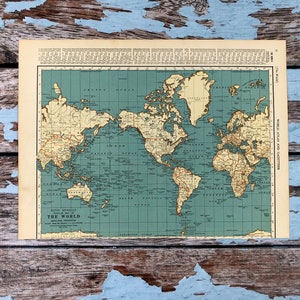 May include: A vintage world map from the Rand McNally World Atlas and Gazetteer. The map features a teal ocean and tan landmasses with detailed geographical information. The map is on a cream-colored paper and is set against a distressed wood background.