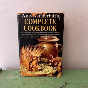 May include: A vintage cookbook titled "Amy Vanderbilt's Complete Cookbook" with a black cover and gold lettering. The cover features a roasted meat dish, a ceramic pot, and text describing the book's contents.