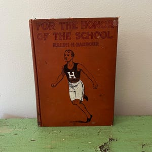 Pode incluir: Um livro antigo intitulado "FOR THE HONOR OF THE SCHOOL" de Ralph H. Barbour. A capa é de cor castanho-avermelhada com uma ilustração de um atleta a correr em preto, branco e castanho. O livro está sobre uma superfície verde.