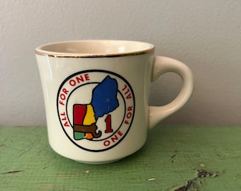 Puede incluir: Taza de cerámica color crema con borde y asa dorados. La taza presenta un gráfico colorido de un contorno estatal con el texto "ALL FOR ONE" que rodea el diseño. También se incluye un número "1" en el diseño.