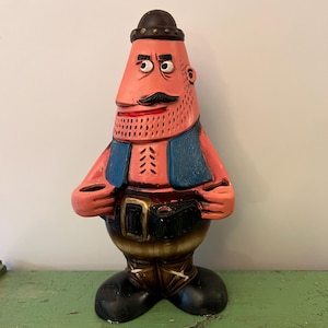 May include: A vintage ceramic figurine of a man with a mustache, wearing a hat, blue vest, and a belt with a gold buckle. The figurine has a pink face and arms, with brown and black accents. The base is brown and black.